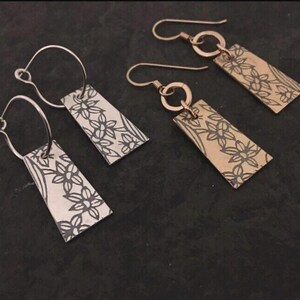 May include: Four pairs of rectangular dangle earrings. Two pairs are silver-toned with silver hooks, and two are gold-toned with gold hooks. Each earring features a floral design in black. The earrings are displayed on a dark surface.