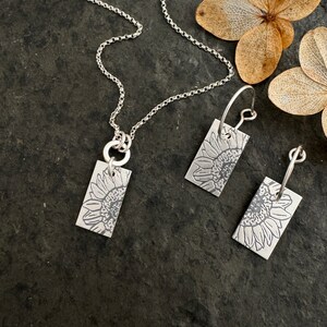 Pode incluir: Um conjunto de colar e brincos de prata com um pingente retangular e brincos com um design de girassol detalhado.