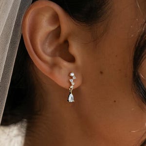 May include: A gold-toned earring featuring a vertical arrangement of three small round clear stones leading to a single dangling pear-shaped clear stone. The earring is worn in the earlobe, complemented by a sheer white veil draped over the hair.