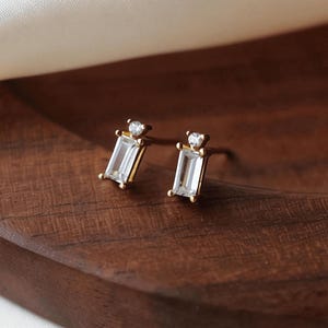 May include: A pair of gold stud earrings. Each earring has a rectangular, clear gemstone set in a gold frame, with a small, round gemstone above. The earrings are presented on a dark wooden surface with white satin fabric.