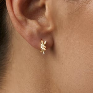 Peut inclure: Une petite boucle d'oreille créole en or présentant un motif de nœud délicat et entrelacé. La surface du nœud est ornée de plusieurs minuscules pierres claires et scintillantes serties le long des bandes métalliques, créant un aspect raffiné.