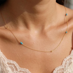 Puede incluir: Un delicado collar de cadena dorada con pequeñas cuentas de turquesa espaciadas uniformemente y diminutas esferas doradas. El collar descansa sobre el cuello, mostrando un diseño sencillo con acentos de piedra azul y cuentas de metal dorado.