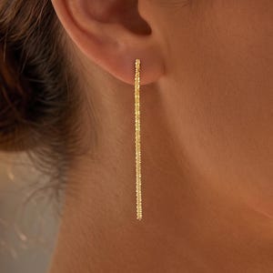 May include: A single gold-colored earring is shown, dangling from an ear. The earring is a long, thin, straight bar with a textured surface. The earring is a bright, shiny gold color.