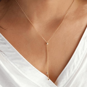 May include: A delicate gold necklace with a Y-shaped design. The necklace features a small sphere at the centre and a dangling chain with two small teardrop-shaped pendants. The necklace is worn against a white shirt.