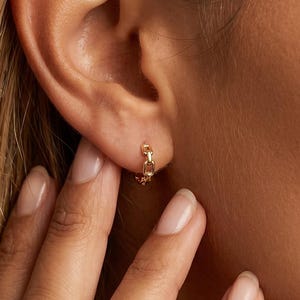 Op de afbeelding: Een kleine gouden oorring met een delicaat schakelkettingontwerp. De oorbel wordt in de oorlel gedragen en toont een gepolijste metalen afwerking en een cirkelvorm die verbonden is met een kort, bungelend kettingsegment met rechthoekige schakels.