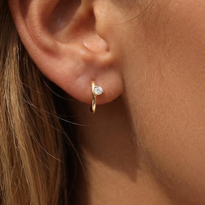 May include: A small gold hoop earring featuring a single, clear, round-cut gemstone set at the front. The earring has a polished metallic finish and a slender, curved design that secures through the earlobe with a simple, minimalist structure.