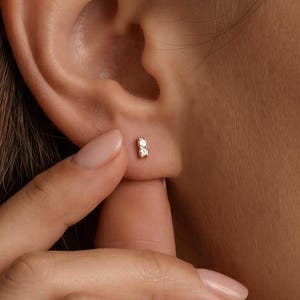 Puede incluir: Un pequeño pendiente de oro con dos diminutas piedras transparentes de corte redondo engastadas verticalmente en una delicada barra de metal. El pendiente está colocado en el lóbulo de la oreja, mostrando un diseño sencillo y pulido.