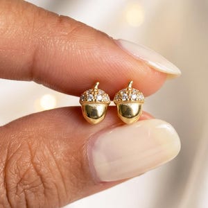 Peut inclure: Une paire de boucles d'oreilles clous en forme de gland, composées d'une base en or lisse et polie et d'un capuchon texturé incrusté de petites pierres claires et scintillantes. Chaque pièce présente une petite tige sur le dessus.