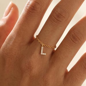 以下が含まれることがあります： ぶら下がるL字型のチャームが付いたゴールドチェーンリング。文字のチャームには、表面に沿って小さな透明な輝く石がセットされています。リングバンドは指に巻き付く繊細なゴールドのリンクで構成されています。