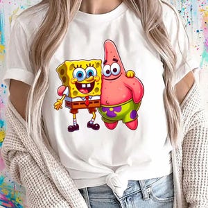 Puede incluir: Camiseta blanca con un gr&aacute;fico de dibujos animados de Bob Esponja y Patricio Estrella. Bob Esponja es amarillo con una corbata roja y pantalones cortos marrones. Patricio es rosa con pantalones cortos verdes. El dise&ntilde;o est&aacute; centrado.