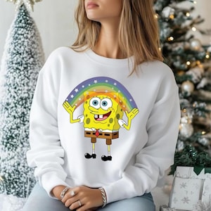 Puede incluir: Sudadera blanca con un gr&aacute;fico colorido de Bob Esponja bajo un arco&iacute;ris. El personaje de dibujos animados es amarillo con corbata y pantal&oacute;n marr&oacute;n. La sudadera es de estilo casual.