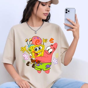 Puede incluir: Camiseta beige con un gr&aacute;fico colorido de Bob Esponja y Patricio Estrella. Bob Esponja es amarillo con una espiral roja en la cabeza, y Patricio es rosa. La camiseta es de estilo casual.