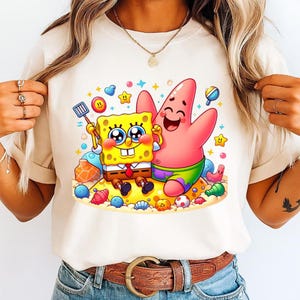 Puede incluir: Camiseta color crema con una colorida ilustraci&oacute;n de dibujos animados de Bob Esponja y Patricio Estrella en una playa arenosa. Bob Esponja sostiene una esp&aacute;tula y Patricio lo abraza. El dise&ntilde;o incluye corazones, estrellas y conchas marinas.