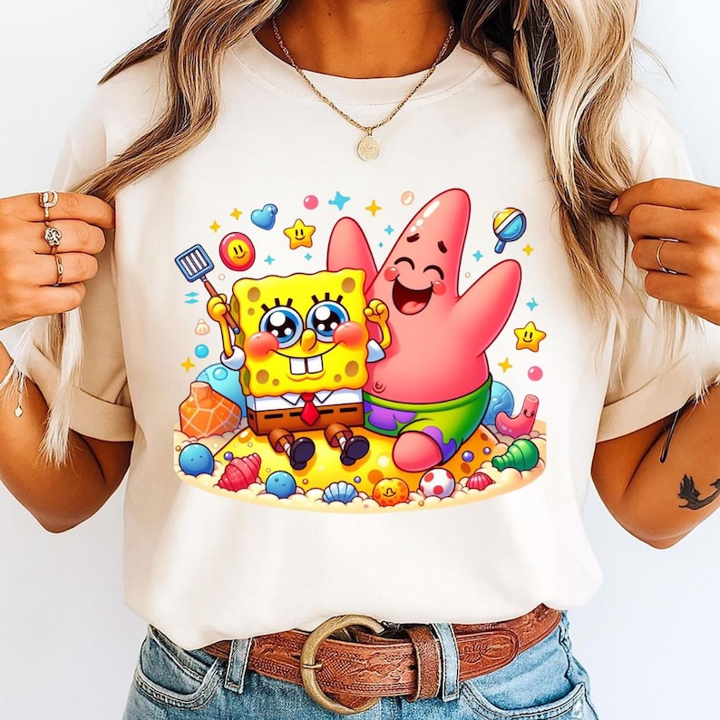 Puede incluir: Camiseta color crema con una colorida ilustraci&oacute;n de dibujos animados de Bob Esponja y Patricio Estrella en una playa arenosa. Bob Esponja sostiene una esp&aacute;tula y Patricio lo abraza. El dise&ntilde;o incluye corazones, estrellas y conchas marinas.
