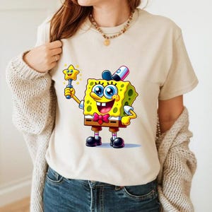 Puede incluir: Camiseta beige con una imagen de dibujos animados de Bob Esponja sosteniendo una varita m&aacute;gica en forma de estrella. Bob Esponja es amarillo con ojos azules, una pajarita roja y un sombrero negro. Manga corta.
