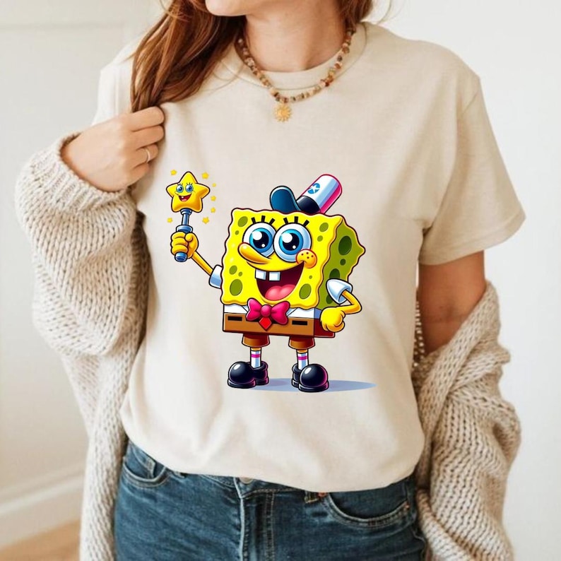 Puede incluir: Camiseta beige con una imagen de dibujos animados de Bob Esponja sosteniendo una varita m&aacute;gica en forma de estrella. Bob Esponja es amarillo con ojos azules, una pajarita roja y un sombrero negro. Manga corta.