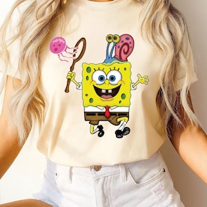 Puede incluir: Camiseta amarilla clara con una imagen de dibujos animados de Bob Esponja sosteniendo una red con una medusa. Bob Esponja sonr&iacute;e, lleva una corbata roja y tiene un caracol en la cabeza. Prenda de vestir informal.