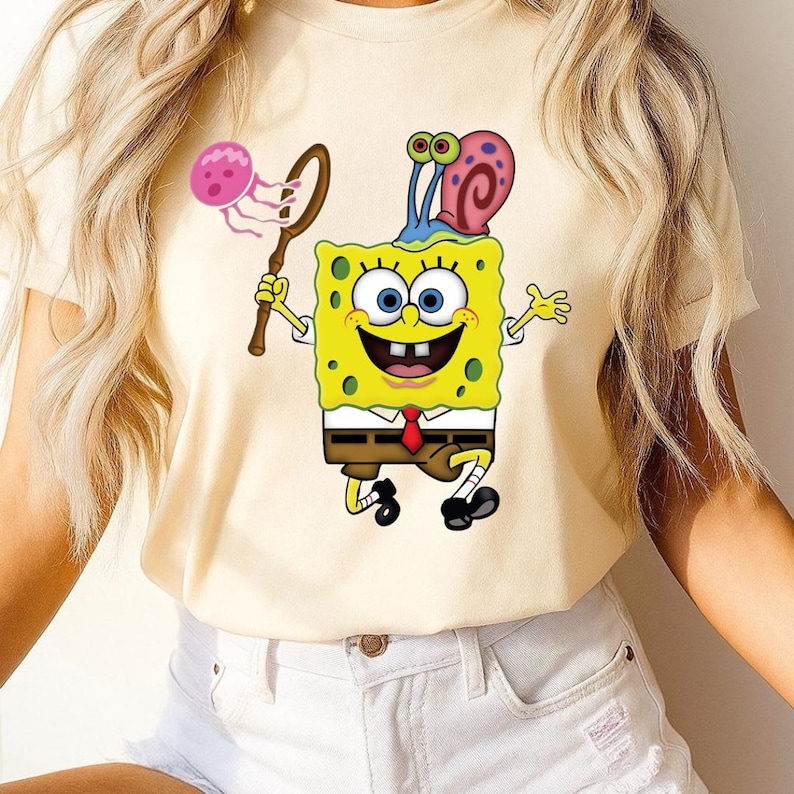 Puede incluir: Camiseta amarilla clara con una imagen de dibujos animados de Bob Esponja sosteniendo una red con una medusa. Bob Esponja sonr&iacute;e, lleva una corbata roja y tiene un caracol en la cabeza. Prenda de vestir informal.