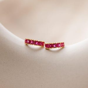 May include: A pair of gold hoop earrings featuring a row of vibrant, square-cut pink gemstones. The earrings have a polished gold metal finish and a curved design, with each stone securely set along the outer edge of the small, delicate hoops.