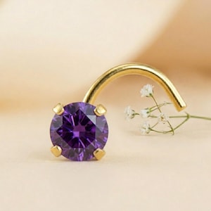 May include: A gold nose ring with a large, round, deep purple gemstone. The gemstone is held in place by four gold prongs. The nose ring has a curved post. A sprig of white flowers is next to the nose ring.
