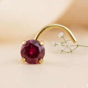 May include: A gold nose ring with a round, faceted, deep red gemstone. The curved post is gold and the gemstone is held in place by four gold prongs. A sprig of white flowers is in the background.