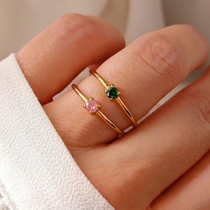 Op de afbeelding: Twee gouden ringen, elk met een enkele edelsteen. Eén ring heeft een roze edelsteen, de andere een groene edelsteen. De ringen zijn dun en delicaat, met een eenvoudig bandontwerp. De edelstenen zijn rond en gezet in een klassieke zetting.