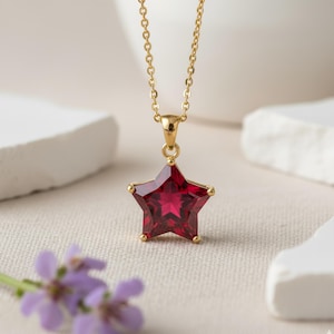 Può includere: Una collana dorata con un ciondolo a forma di stella con una gemma rosso intenso. La stella, larga circa 2.5 cm, è sospesa a una sottile catena dorata. La gemma ha un design sfaccettato.