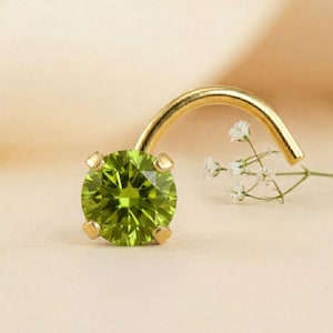 May include: A gold nose stud with a round, faceted, light green peridot gemstone. The gemstone is held by four gold prongs. The curved post is also gold. A sprig of white flowers is in the background.
