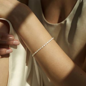 May include: A fine bracelet featuring small, white pearls. The bracelet is displayed on an arm, with the pearls arranged in a single strand. The oval-shaped pearls have a gentle sheen, reflecting the light. A simple and elegant piece of jewellery.