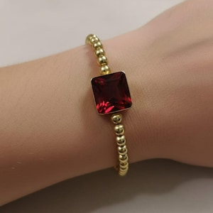 Può includere: Bracciale dorato con perline e una grande gemma rosso scuro quadrata. Il bracciale è indossato su un polso, evidenziando il contrasto tra le perline dorate e la pietra rosso vivo. Un gioiello semplice ma elegante.