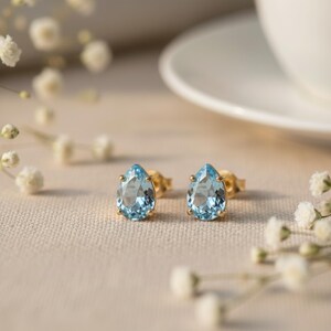 May include: A pair of stud earrings featuring pear-shaped, light blue faceted gemstones set in gold metal prongs. The earrings are positioned side by side, showcasing the clear, brilliant cut of the stones and the polished gold metal settings.