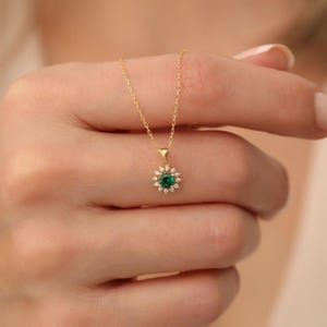 Op de afbeelding: Een goudkleurige ketting met een hanger met een centrale groene edelsteen omringd door kleine, heldere edelstenen. De hanger heeft de vorm van een bloem of zonnestraal. De ketting wordt door een persoon vastgehouden.