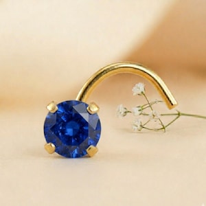 May include: A gold nose stud featuring a round, deep blue gemstone. The gemstone is held in place by four gold prongs. The stud has a curved post for insertion. A sprig of white flowers is in the background.