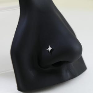 Könnte beinhalten: Ein silberner Nasenpiercing in Form eines vierzackigen Sterns mit einem kleinen, klaren Edelstein in der Mitte. Der Stecker befindet sich auf einer schwarzen Mannequinnase vor weißem Hintergrund.