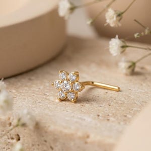 May include: A gold nose stud featuring a flower-shaped cluster of seven clear, round-cut stones. The jewelry piece has an L-shaped post extending from the back of the floral setting, which is composed of one central stone surrounded by six petals.