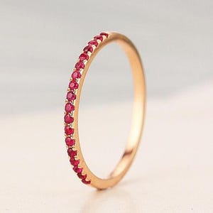 Può includere: Un anello color oro con una fila di piccole pietre preziose rosse. L'anello è sottile e delicato, con le pietre preziose incastonate vicine l'una all'altra nella metà superiore della fascia. L'anello è mostrato su uno sfondo morbido e neutro.