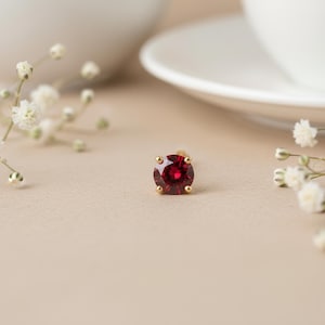 May include: A single gold stud earring with a round, deep red gemstone. The earring is set with four prongs and is displayed on a neutral-toned surface, with white flowers and a blurred background.