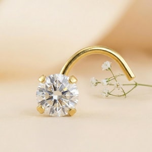 May include: A gold nose ring featuring a round, clear gemstone. The gemstone is held in place by four gold prongs. The curved post is also gold. A sprig of white flowers is in the background.