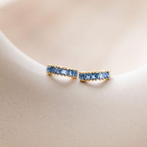 Puede incluir: Un par de pendientes de aro dorados con una hilera de piedras preciosas azules de corte cuadrado. Cada pendiente está diseñado con una banda de metal dorado que sujeta las piedras en un engaste de canal, creando una línea de color continua.