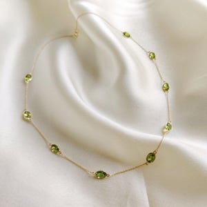 May include: A delicate gold necklace featuring oval-cut peridot gemstones. The necklace has a fine gold chain with evenly spaced, vibrant green peridot stones. The necklace is displayed on a white, silky background.