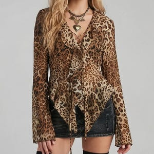 May include: A long-sleeved blouse with a leopard print pattern. The blouse features a ruffled front, a V-neckline, and a tie closure. The model is wearing a necklace with a heart pendant.