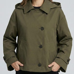 May include: An olive green hooded jacket with a double-breasted design. The jacket features three dark buttons down the front, a collar, and long sleeves with buttoned cuffs. The model is wearing a black shirt underneath.