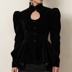May include: A black velvet jacket with a high collar and keyhole detail. The jacket features puff sleeves and a peplum waist. The model is wearing a black skirt with lace trim and black tights. The earrings are black and gold.