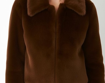 May include: A brown faux fur jacket with a notched collar. The jacket has long sleeves and a straight hemline. The model is wearing black pants. The jacket is a solid color and has a soft, plush texture.