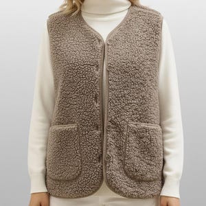 May include: A brown, button-up vest with two front pockets. The vest has a textured, fuzzy appearance. The person in the image is wearing a white turtleneck and white pants. The vest is a neutral color and could be worn in the fall or winter.