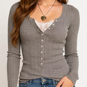 May include: A gray long-sleeve henley shirt with a scoop neck and button-up front. The shirt has a ribbed texture and is paired with blue jeans. A heart-shaped pendant necklace is visible.