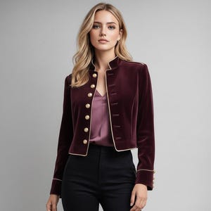 May include: A burgundy velvet jacket with gold buttons and trim. The jacket has a high collar and is worn over a mauve top and black trousers. The jacket's design is reminiscent of a military style.