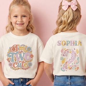 Peut inclure: T-shirt crème avec un motif rétro coloré indiquant "I'm Two Cute" à l'avant. Le dos du t-shirt affiche le nom "Sophia" et un motif floral "2". Le t-shirt est porté par un jeune enfant.