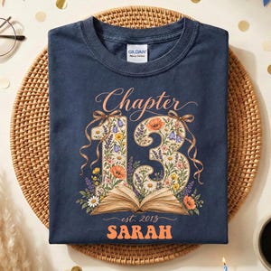 May include: A navy blue t-shirt featuring the text "Chapter 13" above a large number 13 filled with colorful floral illustrations and an open book graphic. Below the book, the text "est. 2013" and the name "SARAH" are printed in orange.