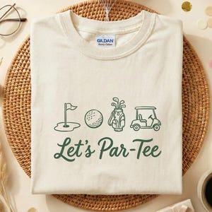 May include: A cream-coloured t-shirt featuring a green golf-themed graphic and the text "Let's Par-Tee." The design includes a golf flag, ball, bag of clubs, and golf cart. The shirt is on a woven mat with birthday decorations.